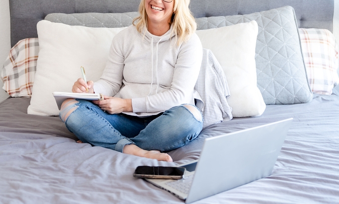 Woman sitting on bed with notebook, pen, laptop, and phone - How to blog consistently for your author business